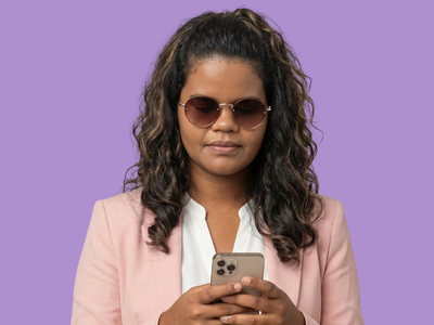 Foto de estúdio de uma mulher de cabelos cacheados e óculos escuros redondos, em pé contra um fundo roxo sólido. Ela veste um blazer rosa claro e segura um smartphone com as duas mãos, olhando para a tela.